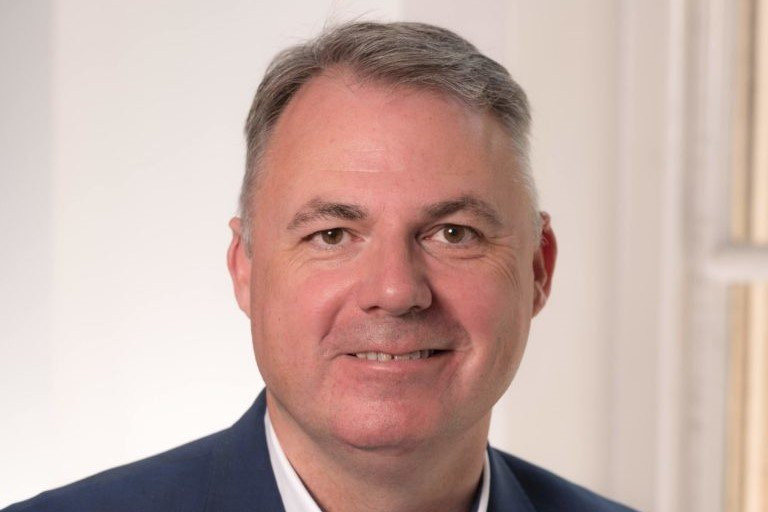 A middle-aged man with short gray hair and a pleasant smile wears a blue blazer. He is positioned against a light-colored, softly blurred background.