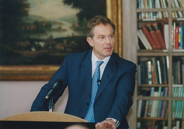 A man in a navy suit and tie stands at a podium, gesturing with one hand while speaking. Behind him is a bookshelf filled with books and a large painting depicting a landscape.