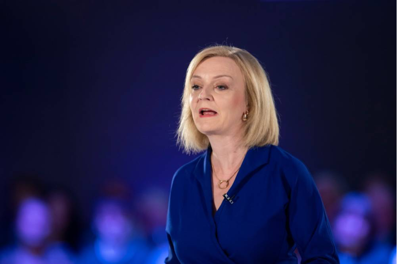 A woman with shoulder-length blonde hair speaks passionately at a podium, wearing a blue blouse. The background features a blurred audience in a dimly lit setting.