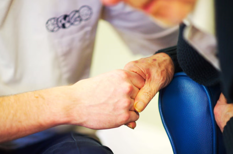 A close-up of two hands clasped together, one hand belonging to a person with visible age spots and a worn appearance, while the other hand appears younger. A blurred figure is partially visible in the background.