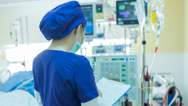 A healthcare professional wearing blue scrubs and a surgical cap checks patient information on a clipboard in a hospital setting, surrounded by medical equipment and monitors.