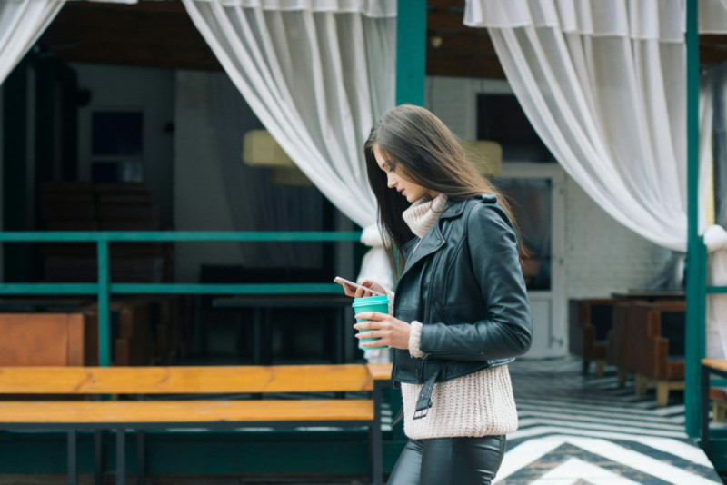 A young woman in a leather jacket and sweater walks outdoors, holding a coffee cup while looking at her phone. The background features a modern, partially covered outdoor area with wooden benches and curtains.