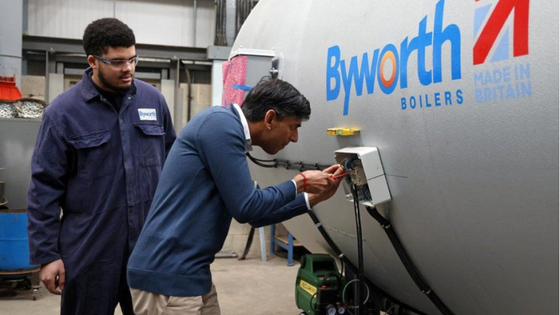 A man in a blue jacket observes another man in a sweater inspecting a control panel on a large boiler marked "Byworth Boilers Made in Britain."