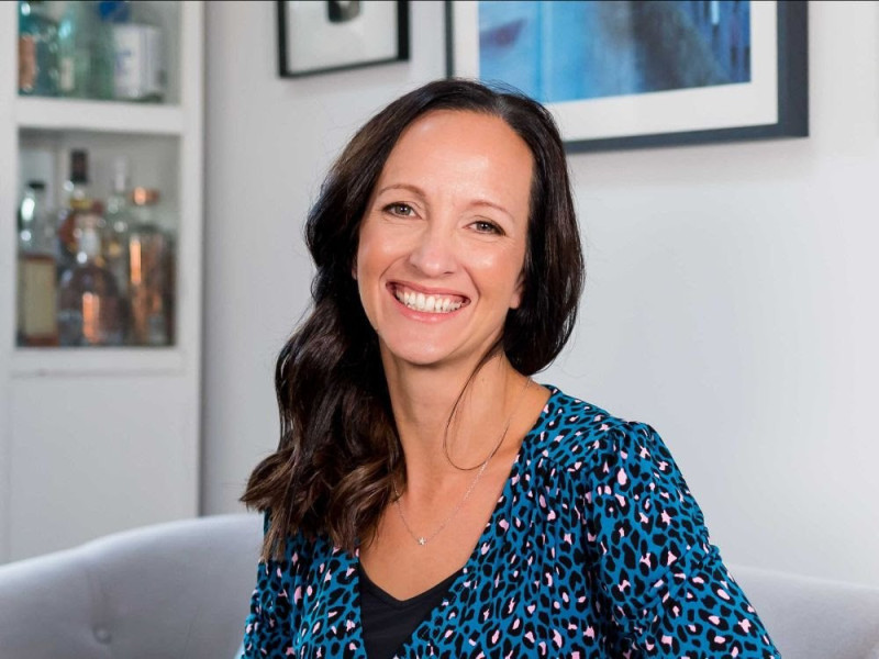 A smiling woman with long, wavy dark hair wears a blue leopard print blouse. She is seated on a light gray couch, with a stylish background featuring shelves and artwork.