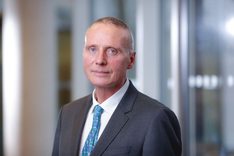 A man with short gray hair and blue eyes, wearing a dark suit and a patterned tie, stands in a professional setting with soft lighting in the background.