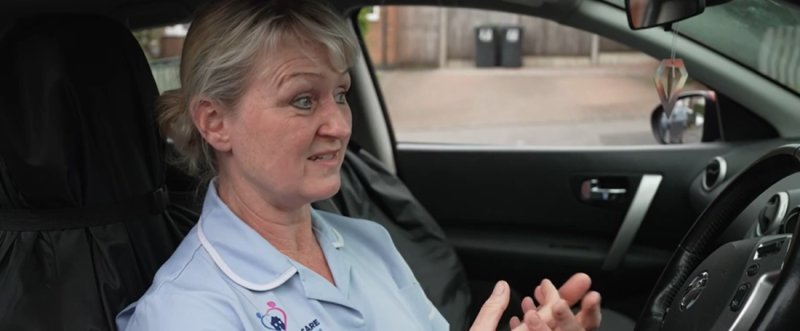 A woman in a light blue uniform sits inside a car, gesturing with her hands while speaking. She has a concerned expression on her face.