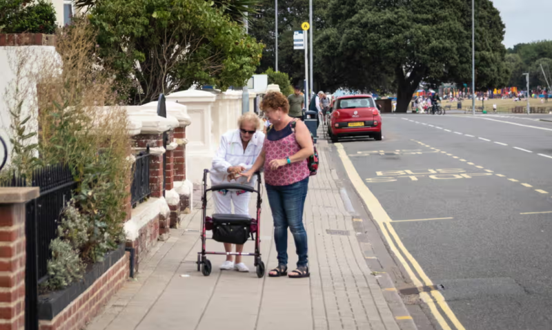 An elderly woman using a walker receives assistance from a younger woman as they walk along a sidewalk. Lush greenery and a road with parked cars are visible in the background.