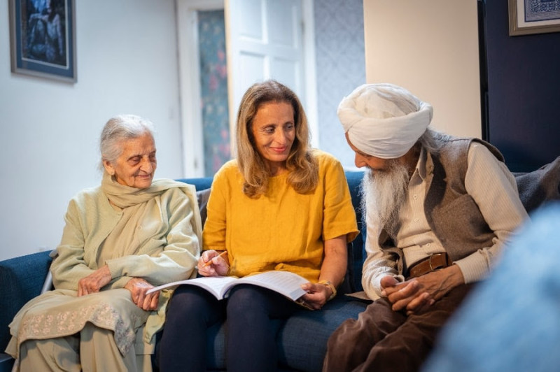 An elderly woman and a man in traditional attire sit on a couch, smiling as they engage in conversation with a younger woman holding a book. The cozy indoor setting features soft lighting and decorated walls.