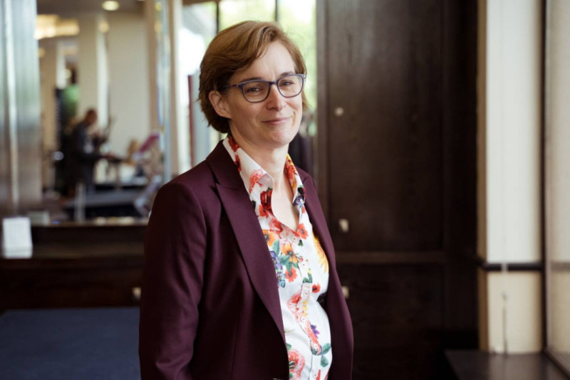 A woman with short hair wearing glasses and a flower-patterned shirt under a maroon blazer smiles warmly while standing indoors in a well-lit environment.