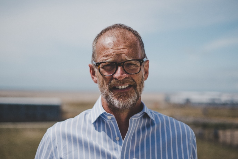 A bearded man with glasses stands outdoors, wearing a blue and white striped shirt, with a blurred landscape and sky in the background.