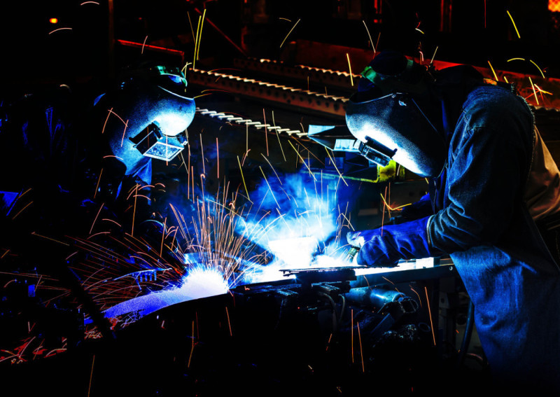 Two welders work on a metal piece, creating bright sparks and blue flames. They are wearing protective gear, including helmets and gloves, set against a dark background.