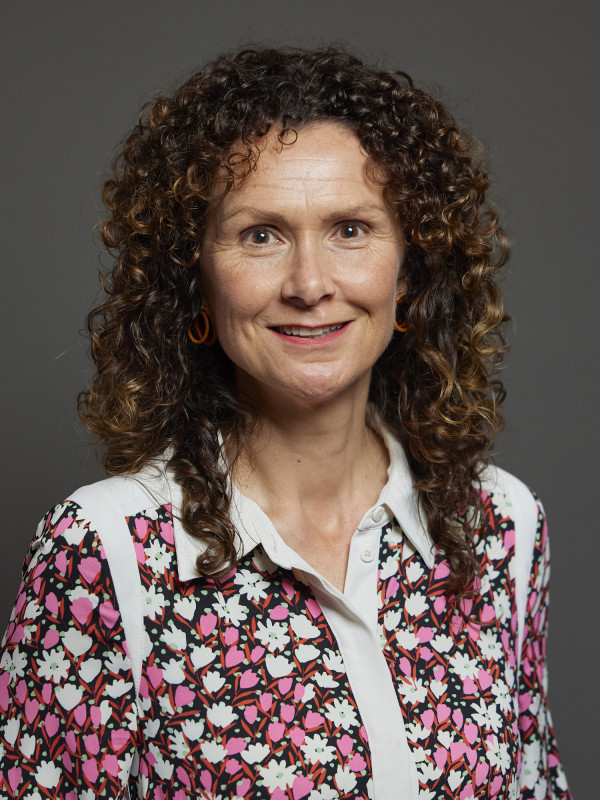A woman with curly brown hair smiles at the camera. She wears a patterned blouse with pink and white flowers and orange circular earrings. The background is gray.