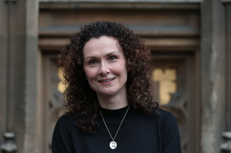 A smiling woman with curly hair poses outdoors in front of a stone structure with intricate details. She wears a black top and a pendant necklace.