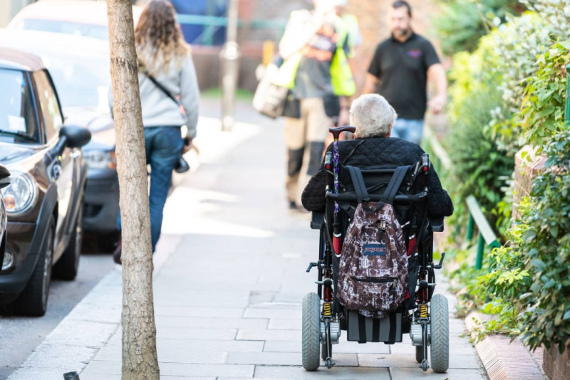 An elderly person in a wheelchair is on a sidewalk, facing away from the camera. A group of workers is walking in the background, with trees and buildings lining the street.