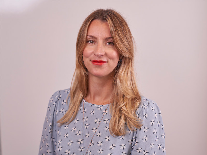 A woman with long, wavy blonde hair smiles at the camera, wearing a blue blouse with a floral pattern and bright red lipstick. The background is a soft, neutral tone.
