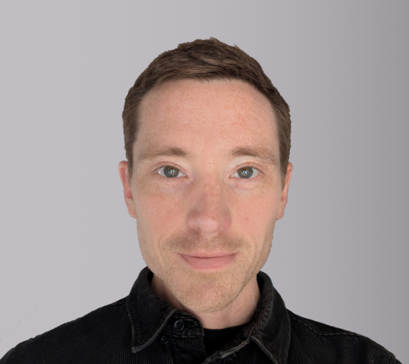 A man with short brown hair and green eyes is looking directly at the camera, wearing a black denim shirt against a light gray background.