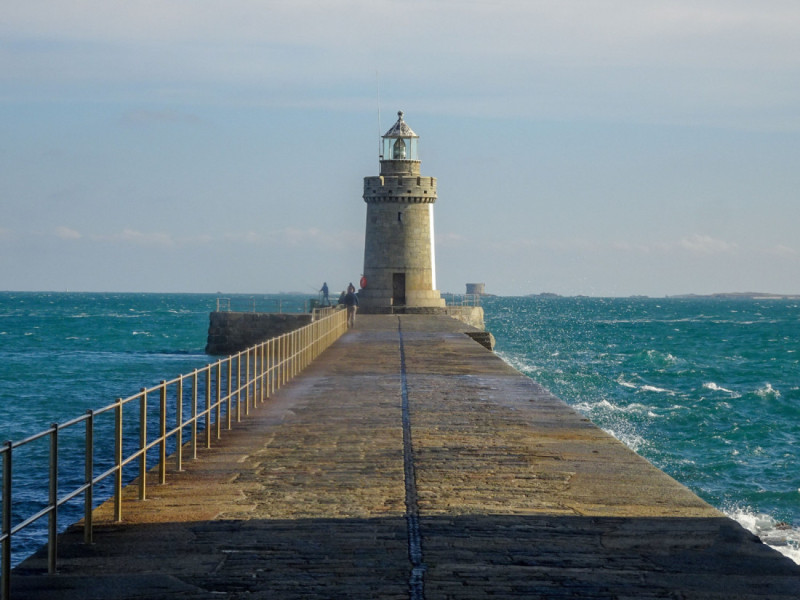 A stone pier stretches into the sea, leading to a tall lighthouse under a partly cloudy sky. Waves crash against the pier's sides, and a few people can be seen walking towards the lighthouse.