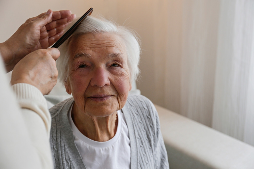 Home Care Marketing - an elderly woman with white hair smiles while someone gently combs her hair. She is seated on a beige sofa with soft natural light coming from a nearby window.