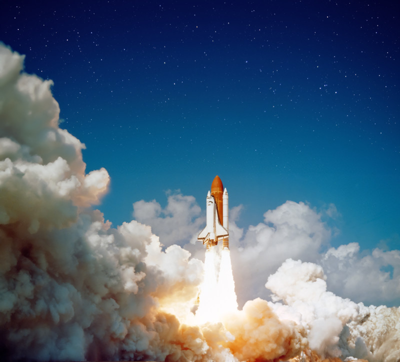 A space shuttle lifts off against a clear blue sky, surrounded by billowing white smoke and clouds. Stars are faintly visible in the background.