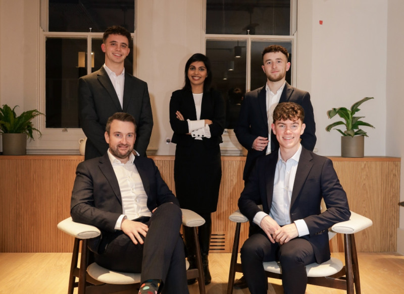 A group of five young professionals in business attire, posing in a stylish office setting. Two men sit on chairs in front, while three stand behind them, all smiling confidently. Potted plants are visible in the background.