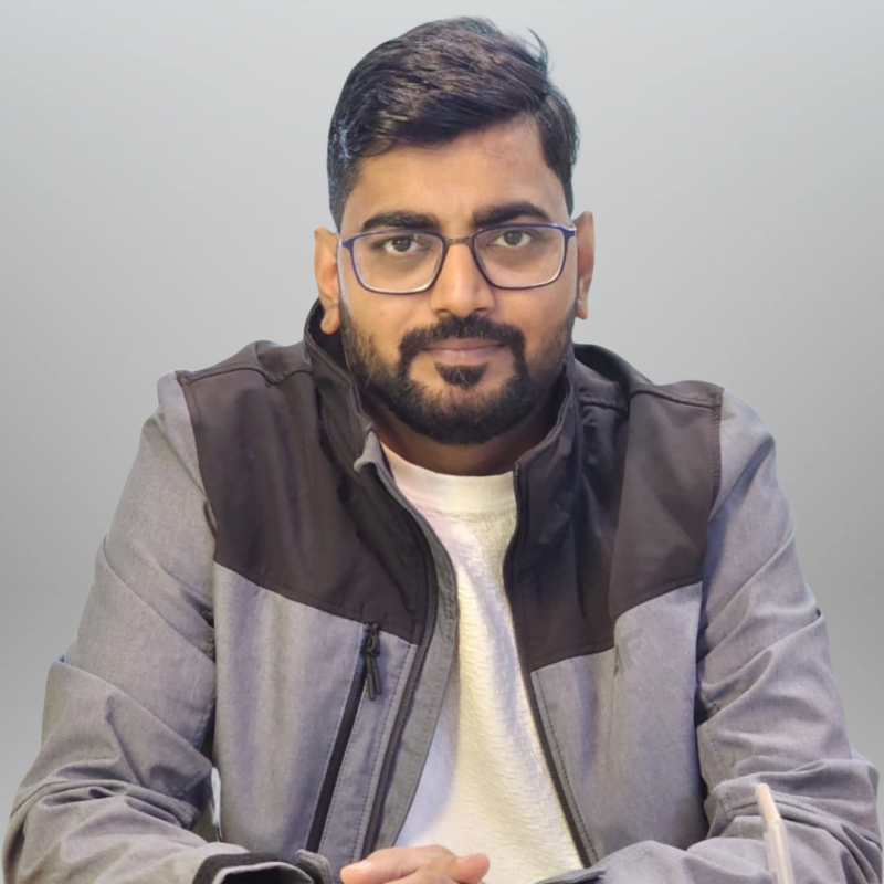 A man with glasses and short black hair sits at a table, wearing a gray jacket with black accents and a white shirt. He has a serious expression and is posed against a light gray background.